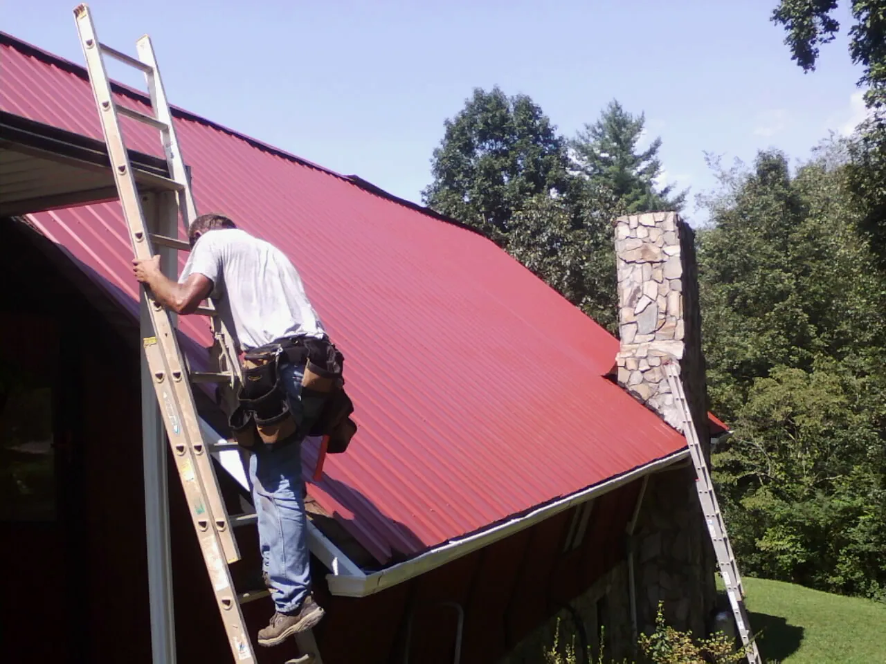 Roofer on a ladder beside a red metal roof and stone chimney — Soffit & Fascia Repair by Roofing Brothers in Canyon